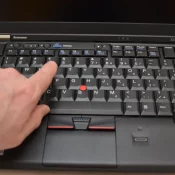 A hand reaching across the keyboard of a ThinkPad X220, with the index finger near the T and 5 keys and the red TrackPoint pointing-stick nub visible just below between the G, H, and B keys. Lenovo logo on the left of the palm rest, ThinkPad logo on the right, X220 model name on the bezel.